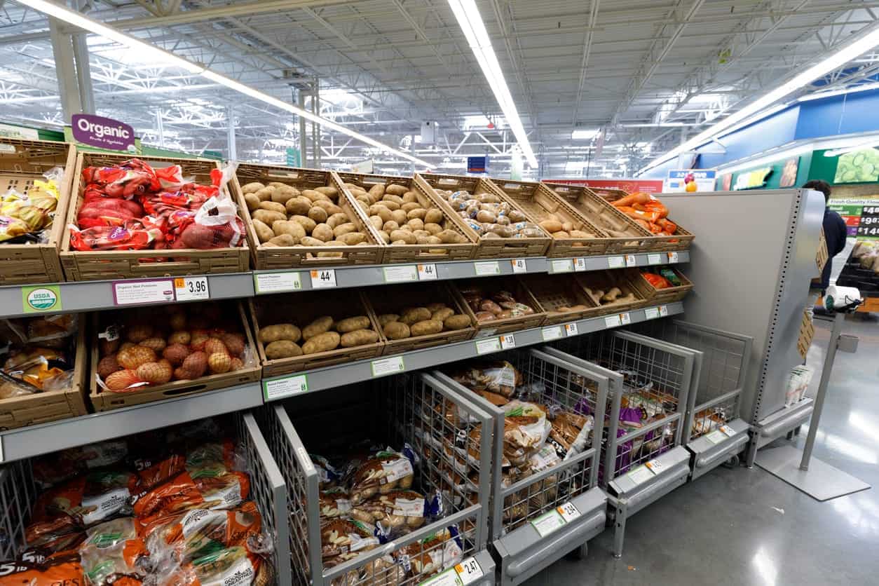 Supermarket produce aisle with potatoes, carrots, and onions on display.