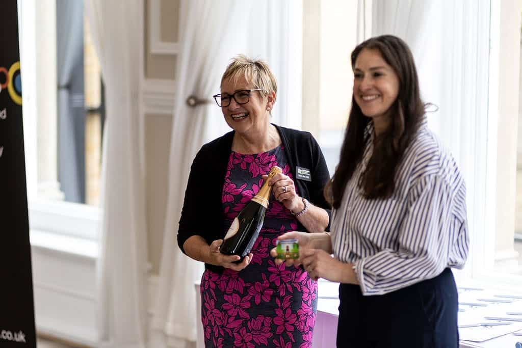 Two women exchanging a bottle of champagne at a networking event.